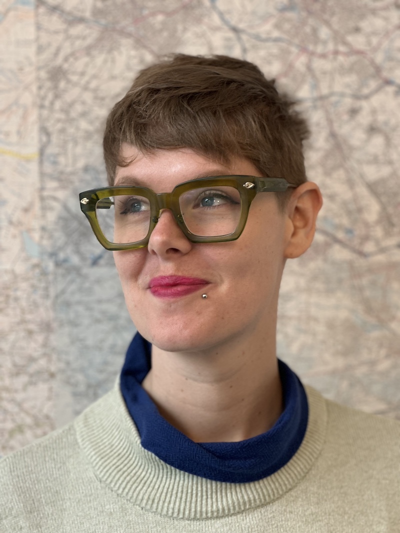 Heidi Swigon is infront of a map with light brown hair, funky chunky broawn glasses and a cream coloured jumper.
